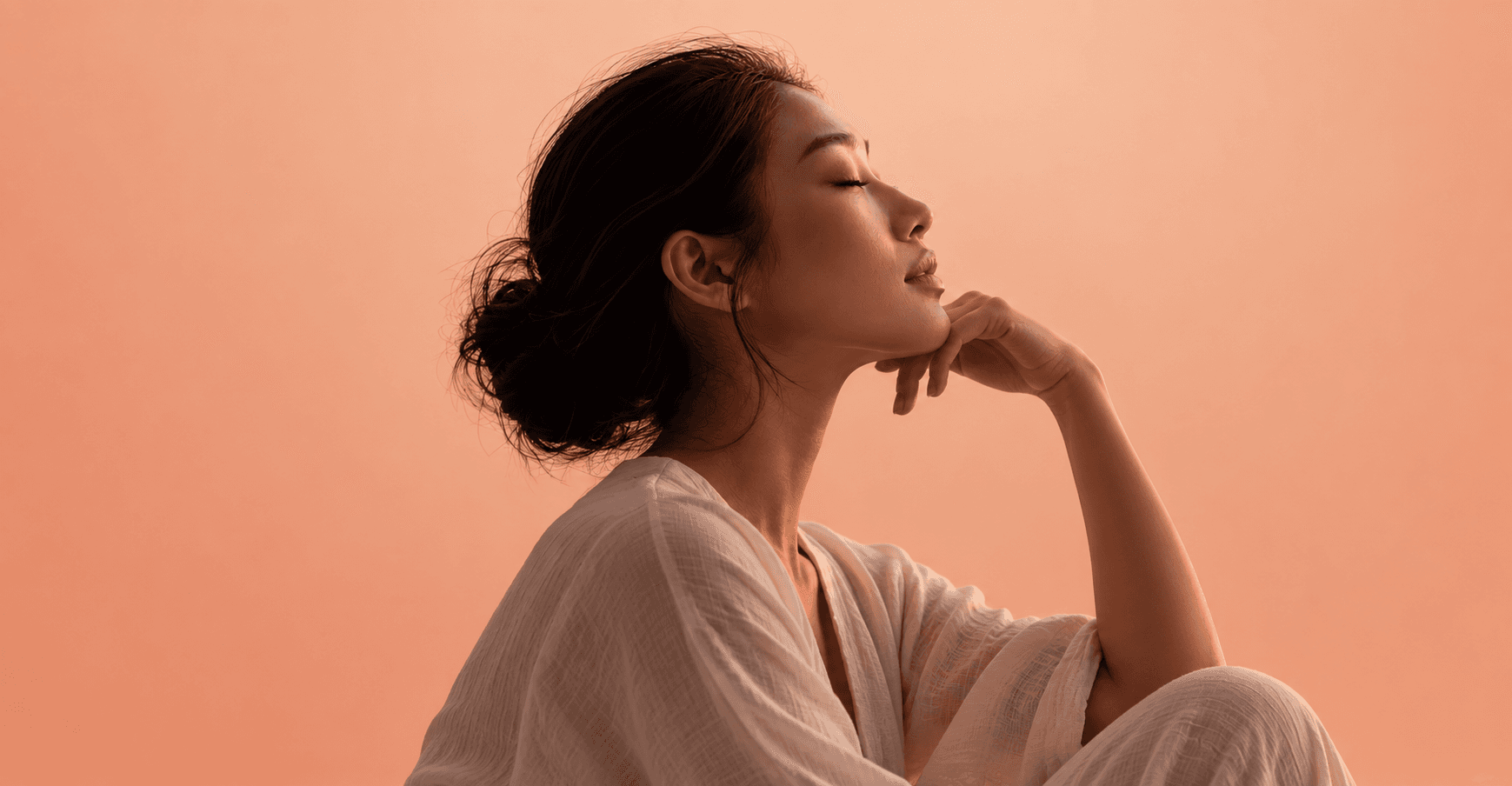 Woman with her eyes closed, resting her chin on her hand against a peach background