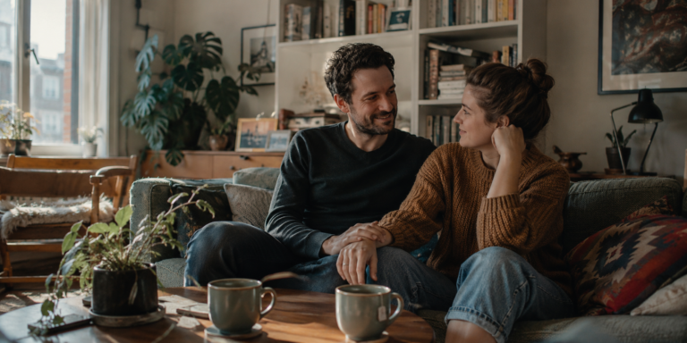 A couple sitting close together on a sofa in a sunlit living room with houseplants and two mugs of tea on a wooden table